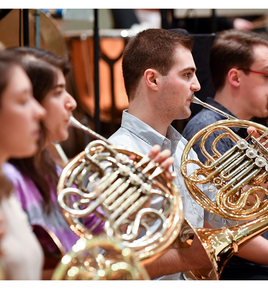 A group of students, performing and holding a French horn in a orchestra rehearsal.